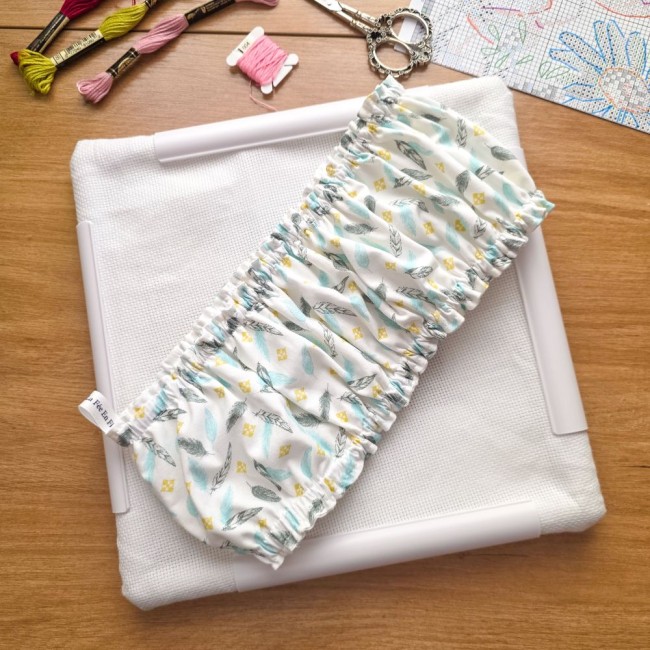 grime-guard-sur-cadre-qsnap-blanc-plumes-pastel-angelique-la-fee-en-fil Un couvre-cadre (grime guard) élastiqué en coton blanc imprimé de plumes pastel bleues et grises et de petits losanges jaunes, posé en diagonale sur une toile Aïda blanche fixée sur un cadre carré blanc de type Q-Snap. L'étiquette de la marque est visible