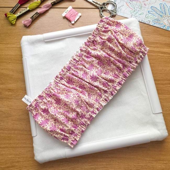Vue rapprochée d'un grime guard housse protège-toile modèle Samba en tissu coton à motifs floraux rose et violet installé sur un cadre carré Q-Snap, avec l'étiquette de la marque visible, échevettes de fils et ciseaux vintage en arrière-plan, création art