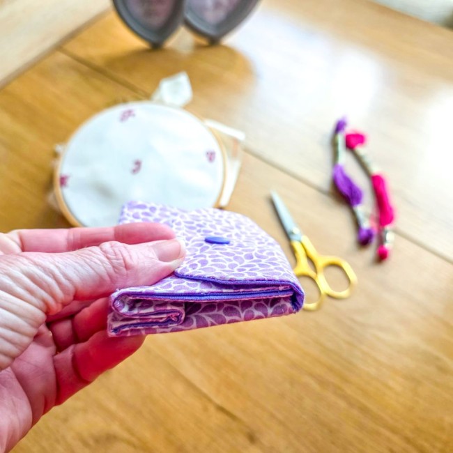 Gros plan sur la prise en main d'une mini poubelle à fils de table pliante fermée à plat modèle Lavande en tissu coton violet à motifs feuillage, avec tambour à broder, ciseaux dorés et échevettes de fils en arrière-plan, création artisanale Angélique La 