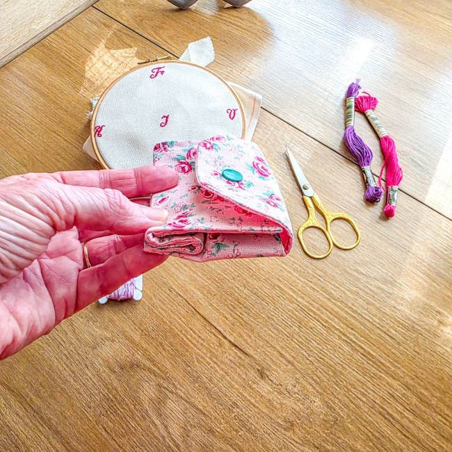 Mini poubelle à fils nomade pliable tenue en main fermée modèle Romane, en tissu rose à motif de roses rouges, montrant son format compact à côté d'un tambour à broder et de ciseaux dorés, création artisanale Angélique La Fée En Fil.