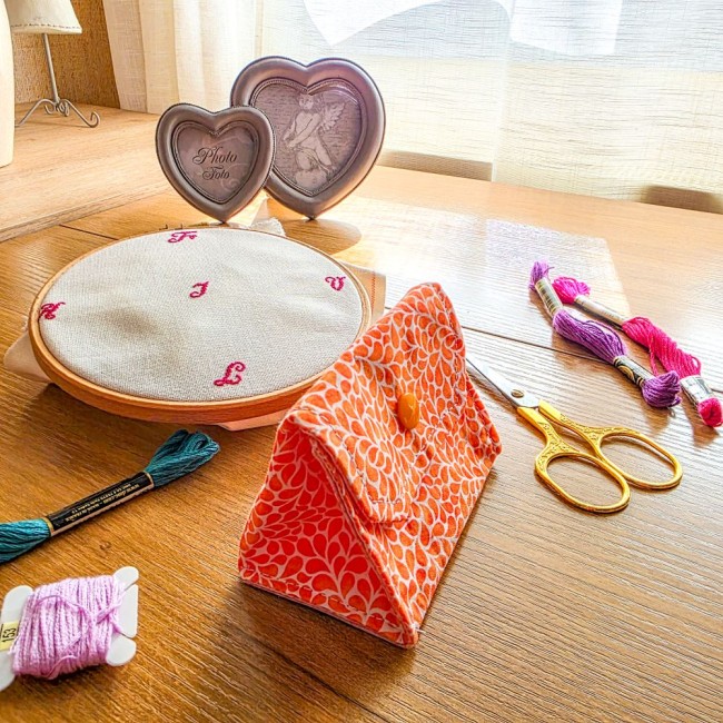 Vue de trois quarts d'une mini poubelle à fils de table pliante modèle Mandarine en tissu coton orange à motifs gouttelettes blanches ouverte sur une table, avec un tambour à broder, des ciseaux dorés et des échevettes de fils, création artisanale Angéliq