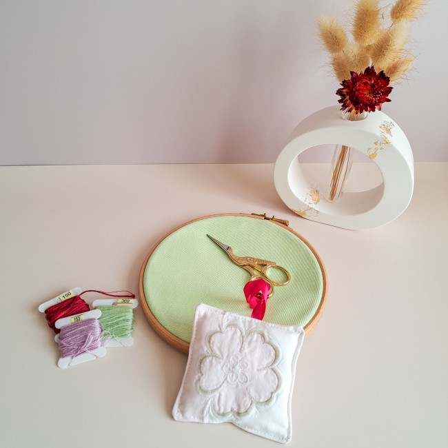 bijou-ciseaux-brodeuse-fleur-blanche-ruban-rose-tambour-broderie-angelique-la-fee-en-fil Bijou de ciseaux pour brodeuse en forme de coussinet blanc brodé d’une fleur, attaché à une paire de ciseaux dorés avec un ruban rose, posé sur un tambour de broderie vert avec fils à broder et décoration florale, création Angélique La Fée En Fil.