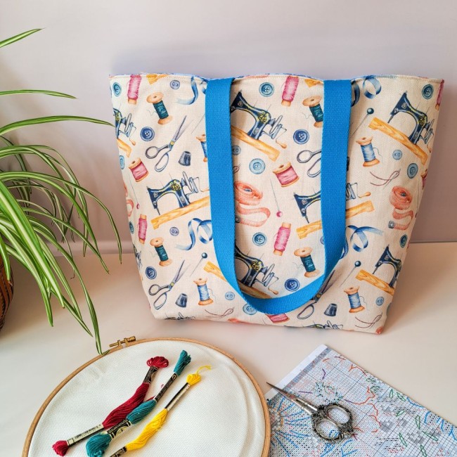 posé sur une table, un sac cabas ouvert devant un tambour à broder, des echevettes de fil à broder, un ciseau à broder et un diagramme de point de croix. Création Angélique La Fée En Fil  . Création Angélique La Fée En Fil