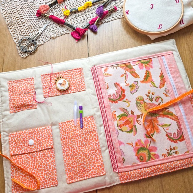 Vue intérieure de la trousse de brodeuse Savannah avec pochette zippée en vinyle transparent révélant le tissu floral aux motifs d'oiseaux, poches en tissu orange et pique-aiguille intégrée, création artisanale Angélique La Fée En Fil.