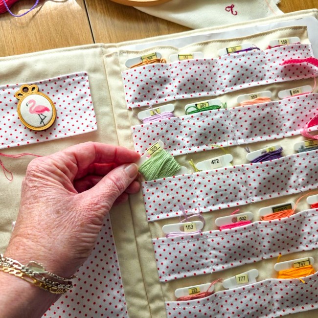 trousse-brodeuse-rangement-cartonnettes-fils-a-broder-alice-angelique-la-fee-en-fil Main attrapant une cartonnette de fil à broder dans les compartiments de la trousse de brodeuse Alice à pois rouges, création artisanale Angélique La Fée En Fil.