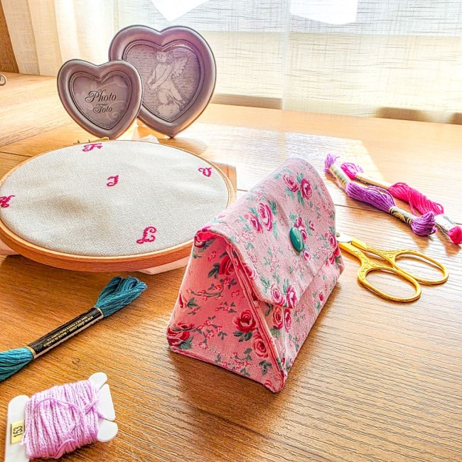 Vue de trois-quarts d'une mini poubelle à fils pliable fermée modèle Romane, en tissu rose à motif de roses rouges avec rabat et bouton-pression vert, posée sur une table en bois à côté d'un tambour à broder, de ciseaux dorés et d'échevettes, création art