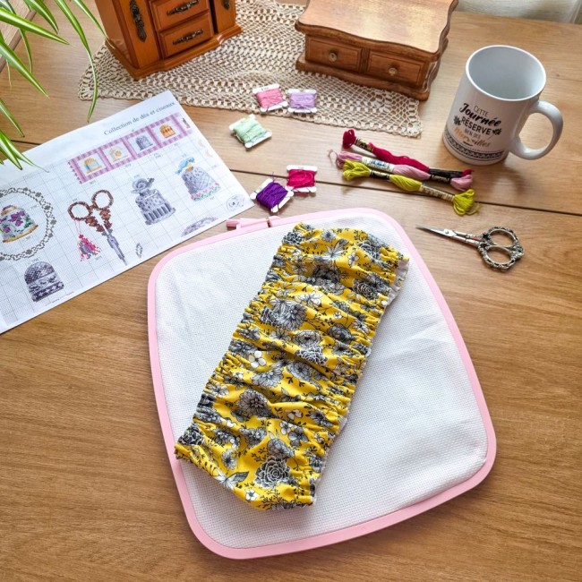 Grime guard modèle Julie en tissu jaune floral plissé, posé au centre d'un tambour à broder rectangulaire Nurge rose avec sa toile à broder sur une table en bois par Angélique La Fée en Fil.