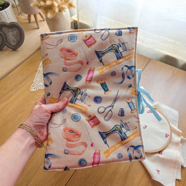 trousse-brodeuse-fermee-mercerie-vintage-mains-et-merveilles-angelique-la-fee-en-fil Main tenant la trousse de brodeuse Mains et Merveilles fermée, montrant la couverture en tissu aux motifs mercerie vintage et son lien en ruban bleu ciel, création artisanale Angélique La Fée En Fil.
