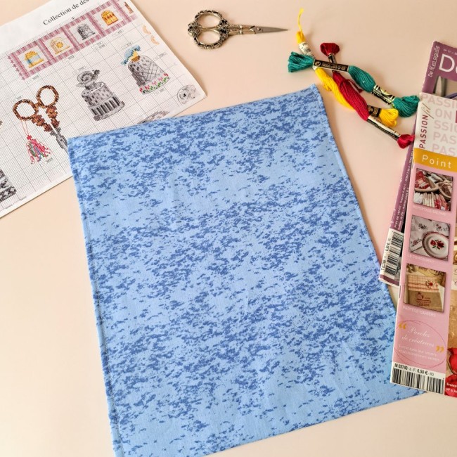 Posé sur la table, une pochette à projet retourné de couleur bleu. A côté on y trouve des ciseaux à broder, des échevettes, une grille de point compté et des magazines de point de croix. Une création Angélique La Fée En Fil