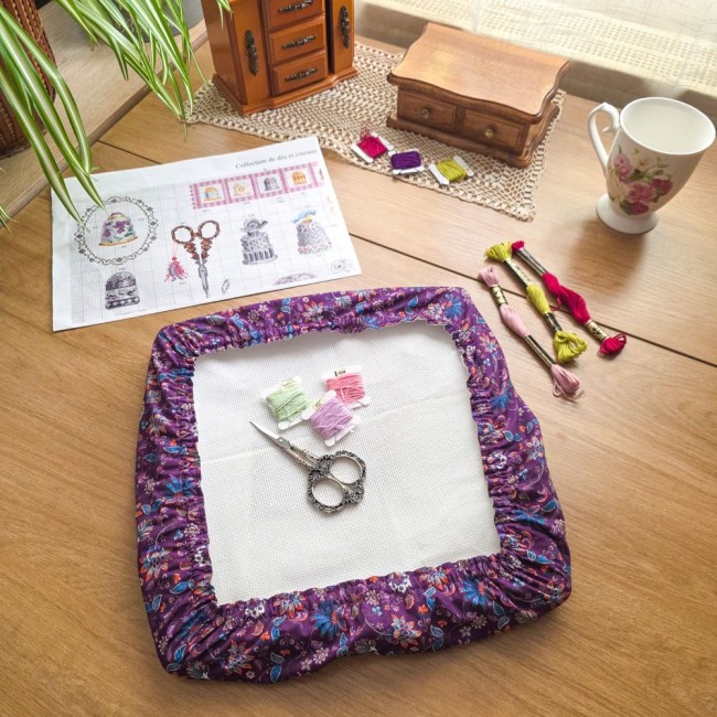 Vue de dessus d'un grime guard modèle Cachemire en tissu violet aux motifs floraux et cachemire multicolores, installé sur un cadre Q-Snap carré avec ciseaux de brodeuse, échevettes de fils et grille de point de croix, création artisanale Angélique La Fée