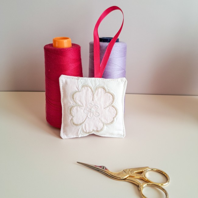 bijou-ciseaux-brodeuse-fleur-blanche-ruban-rose-angelique-la-fee-en-fil Bijou de ciseaux pour brodeuse en forme de coussinet blanc brodé d’une fleur, orné d’un ruban rose, présenté avec des bobines de fil rouge et violet et une paire de ciseaux dorés, création Angélique La Fée En Fil.