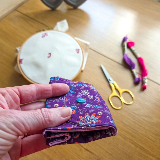 Gros plan sur la finesse et la prise en main de la mini poubelle à fils nomade modèle Cachemire en tissu violet aux motifs floraux et cachemire, repliée à plat avec bouton-pression pour un rangement facile dans une trousse de brodeuse ou pochette à projet