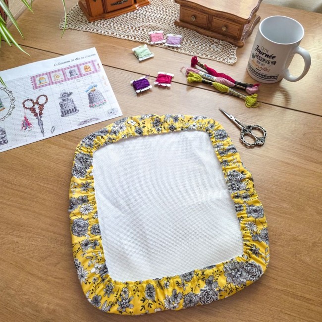 Protège-toile grime guard rectangulaire modèle Julie en tissu jaune floral entourant un métier nurge, posé à plat sur un bureau avec matériel de broderie par Angélique La Fée en Fil.