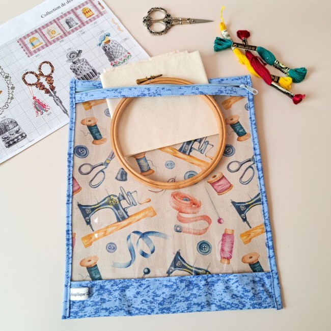 posé sur la table un sac à projet bleu avec un imprimé couture. A l'intérieur il y a un tambour à broder et une toile à broder. A côté une grille de point compté, des ciseaux à broder et des echevettes. Création Angélique La Fée En Fil 