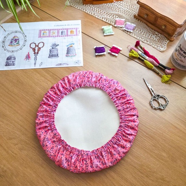 grime-guard-sous-bois-rose-tambour-rond-broderie-angelique-la-fee-en-fil Un couvre-tambour (grime guard) rond modèle Sous-bois Rosé en tissu de coton vieux rose aux motifs de petites branches et feuillages violet prune. Il enserre une toile Aïda blanche tendue sur un cercle à broder rond. La mise en scène sur une table en bois