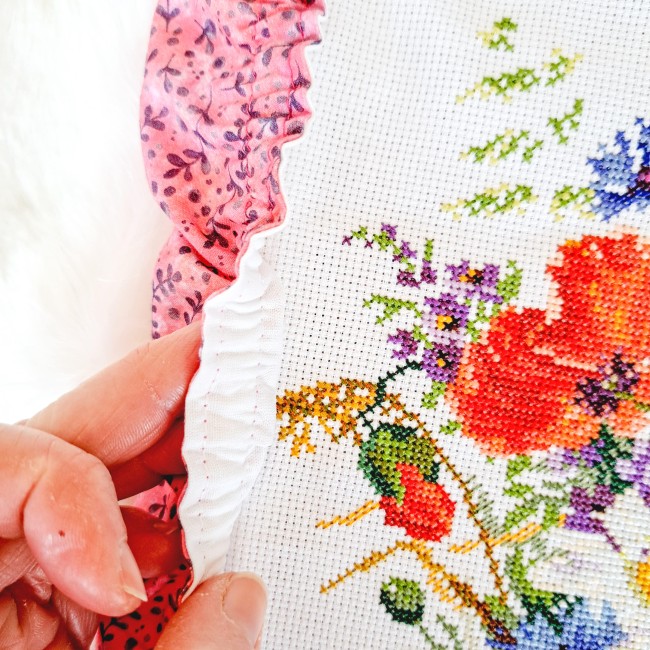 Gros plan d'une main soulevant le bord élastique blanc d'un grime guard modèle Sous-Bois Doré en tissu vieu rose à motifs de feuillage mauve sur une toile brodée au point de croix par Angélique La Fée en Fil