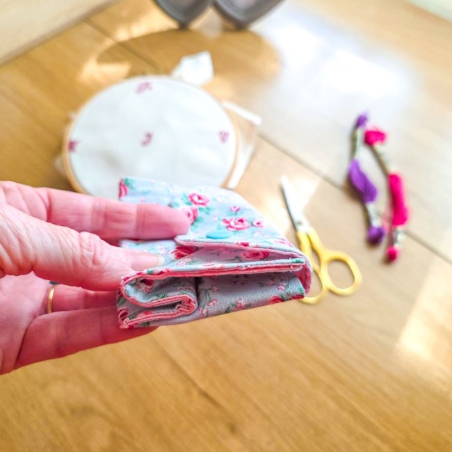 Gros plan de la mini poubelle à fils pliable fermée modèle Bianca tenue en main, montrant sa finesse et sa taille compacte en tissu gris clair aux motifs bucoliques de roses rouges, avec tambour à broder, ciseaux dorés et échevettes en arrière-plan, créat