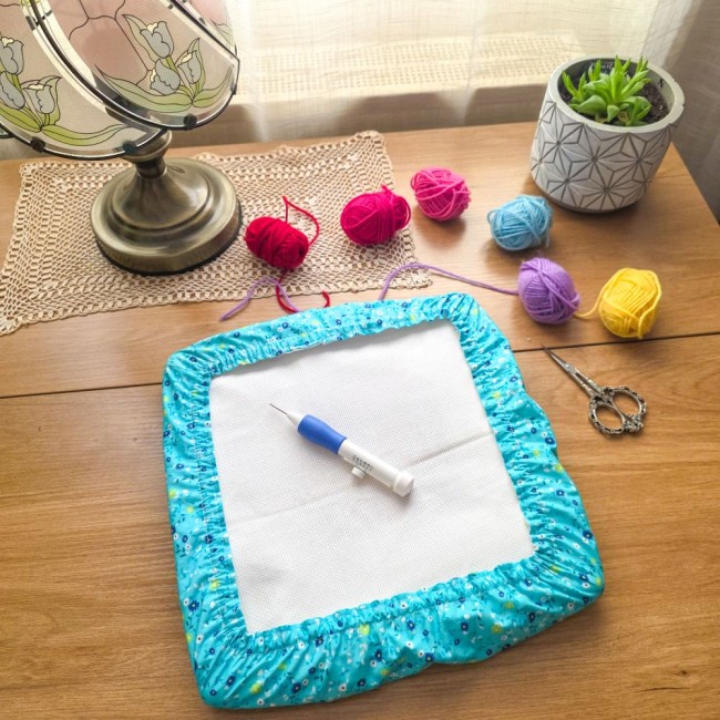 grime-guard-punch-needle-bleu-turquoise-fleuri-angelique-la-fee-en-fil Un grime guard (couvre-cadre) rectangulaire en coton bleu turquoise imprimé de petites fleurs blanches et jaunes, protégeant une toile Aïda. Une aiguille magique à punch needle bleue et blanche est posée au centre. Décoration créative sur table en bois av