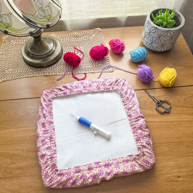 Vue d'ensemble d'un grime guard housse protège-toile modèle Samba en tissu coton à motifs floraux rose et violet, ajusté sur un cadre Q-Snap carré avec un stylo punch needle au centre, pelotes de laine colorées et ciseaux vintage en arrière-plan, création