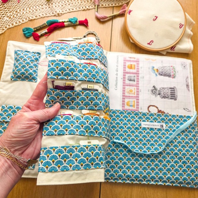 trousse-brodeuse-rangement-cartonnettes-fil-a-broder-grille-mains-et-merveilles-angelique-la-fee-en-fil Main soulevant la page rangement cartonnettes de la trousse de brodeuse Mains et Merveilles aux motifs éventails bleus, dévoilant l'emplacement pour grille de broderie, création artisanale Angélique La Fée En Fil.
