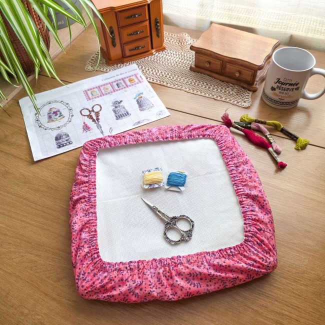 protection-toile-broder-rose-feuille-qsnap-angelique-la-fee-en-fil Un grime guard (couvre-cadre) carré modèle Sous-bois Rosé en coton vieux rose orné de feuillages violets, installé sur un cadre Q-Snap avec sa toile blanche. L'ouvrage est posé sur une table en bois clair. La mise en scène comprend deux bobines de fil (ja