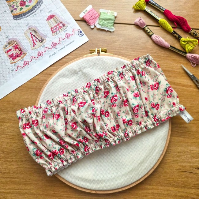 Vue d'ensemble d'un grime guard housse protège-toile modèle Vanille posé en travers sur un tambour à broder rond en bois, en tissu écru vanille aux motifs bucoliques de roses roses et rouges avec étiquette de marque, entouré d'une grille de broderie, éche