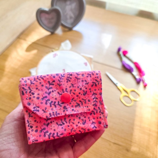 Vue de face d'une mini poubelle à fils pliable fermée modèle Sandy Rose tenue en main, montrant le rabat et le bouton-pression rose sur tissu rose aux motifs de feuillage violet, avec ciseaux dorés et tambour à broder en arrière-plan, création artisanale 