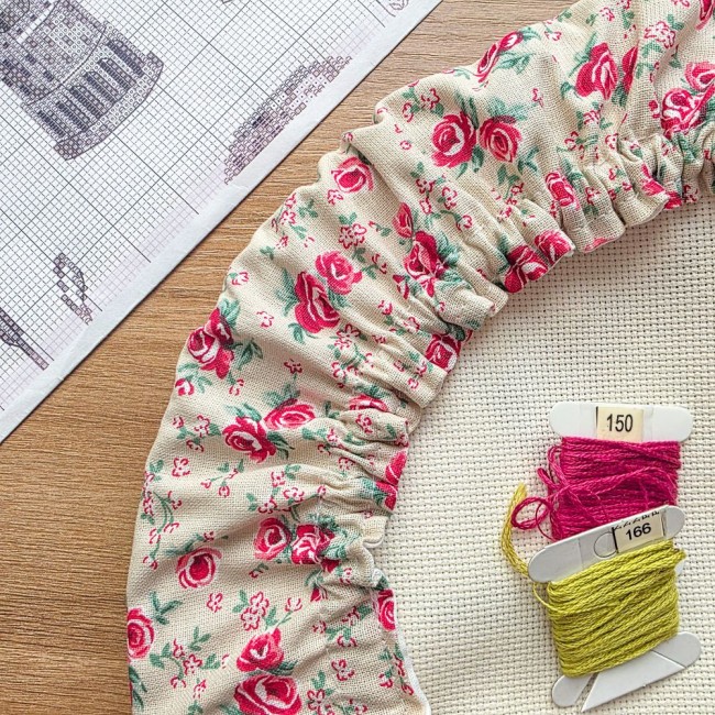 Gros plan sur les fronces et le tissu d'un grime guard modèle Vanille en tissu écru vanille aux motifs bucoliques de roses roses et rouges, posé sur une toile Aïda avec cartes de fils et grille de broderie en arrière-plan, création artisanale Angélique La