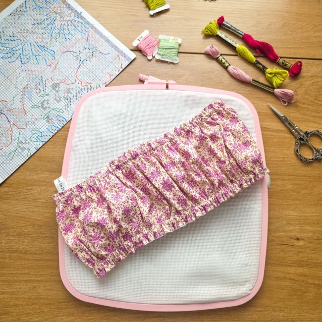 Vue d'ensemble d'un grime guard housse protège-toile modèle Samba en tissu coton à motifs floraux rose et violet, ajusté en diagonale sur un cadre rectangulaire rose de type Nurge , avec grille de broderie, échevettes de fils et ciseaux vintage en arrière