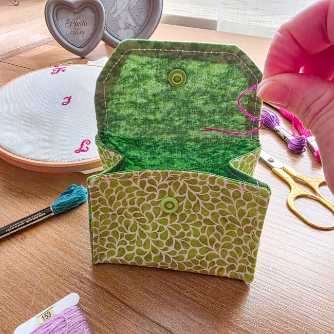 Gros plan d'une main déposant une chute de fil rose dans la poubelle à fils de table ouverte modèle Verveine, doublée d'un tissu vert foncé, avec tambour à broder, ciseaux dorés et échevettes de fils en arrière-plan, création artisanale Angélique La Fée E