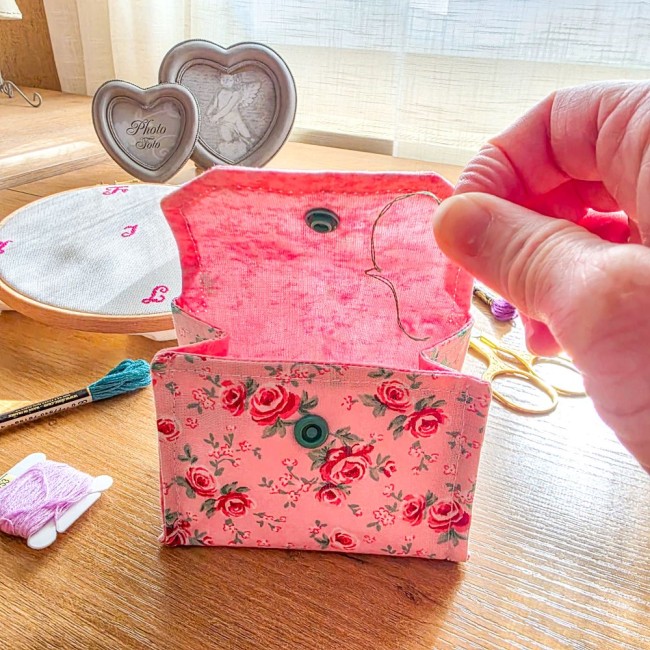Mini poubelle à fils pliable ouverte modèle Romane en tissu rose imprimé de roses rouges et doublure rose, montrant une main y déposant un brin de fil, posée sur une table en bois avec du matériel de broderie, création artisanale Angélique La Fée En Fil.