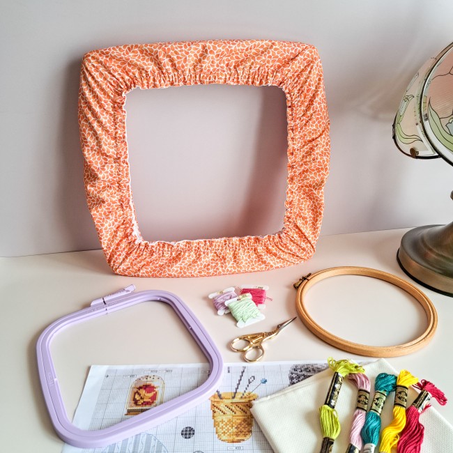 Grime Guard carré en tissu couleur mandarine à motifs, issu de la collection 'Lumière Végétale', installé sur un cadre Q-Snap. Il est entouré d'accessoires de broderie : fils moulinés, ciseaux dorés, tambour rond en bois, nurge, toile Aïda et diagramme.