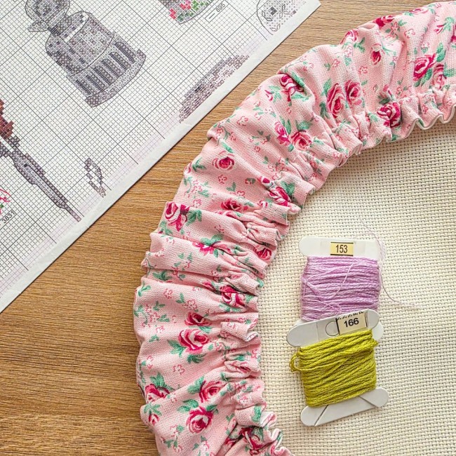 grime-guard-gros-plan-details-tissu-romane-angelique-la-fee-en-fil Gros plan sur les fronces et le tissu d'un grime guard modèle Romane en tissu rose aux motifs bucoliques de fleurs et roses, posé sur une toile Aïda avec cartes de fils et grille de broderie en arrière-plan, création artisanale Angélique La Fée En Fil.