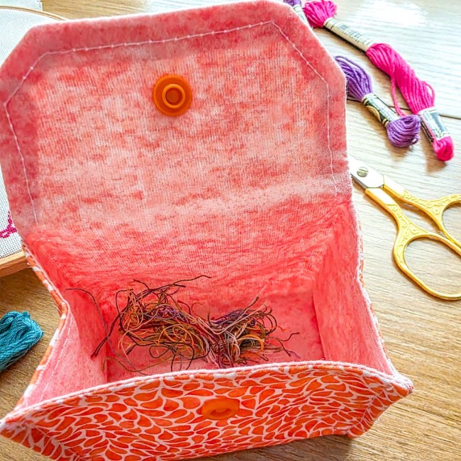 Gros plan sur l'intérieur d'une mini poubelle à fils de table pliante modèle Mandarine en tissu coton orange, garnie de chutes de fils de broderie colorés, avec doublure rose unie, bouton-pression et ciseaux dorés à l'arrière-plan, création artisanale Ang