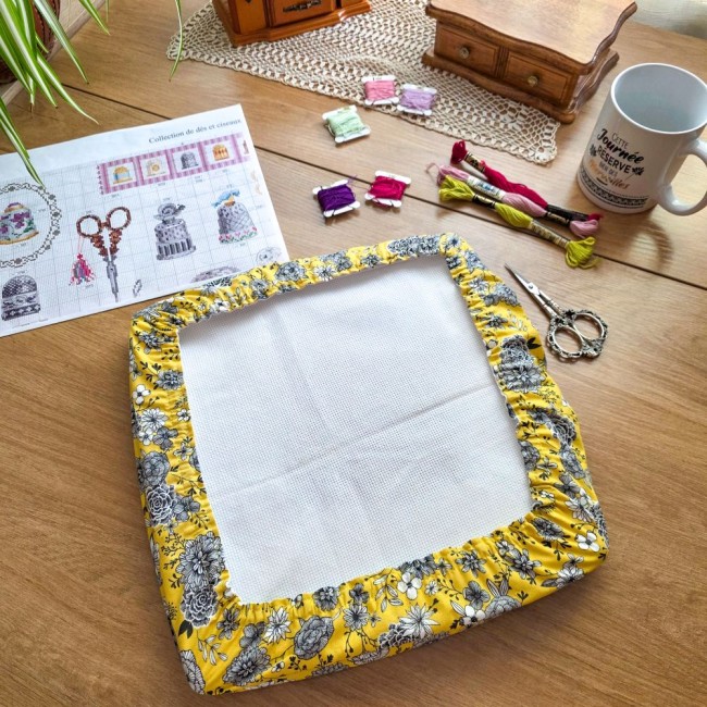 Vue d'ensemble d'un grime guard modèle Julie imprimé de fleurs sur fond jaune protégeant un métier à broder, q-snap, posé sur une table en bois avec accessoires de broderie par Angélique La Fée en Fil.