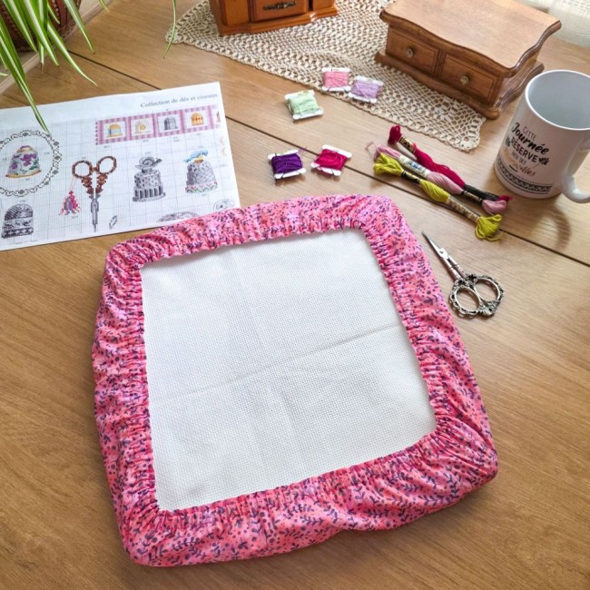 grime-guard-sous-bois-rose-cadre-qsnap-broderie-angelique-la-fee-en-fil Un couvre-cadre (grime guard) carré de type Q-Snap modèle Sous-bois Rosé en coton vieux rose à motifs de délicates branches et feuillages violet prune. Il enserre une toile Aïda blanche tendue. La mise en scène sur une table en bois comprend un diagramme