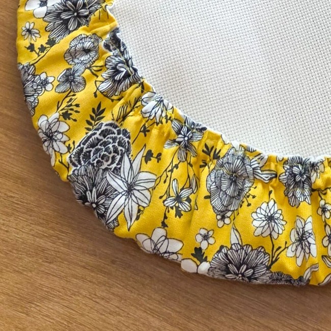 Gros plan sur les détails de couture du grime guard modèle Julie en tissu jaune aux motifs floraux noirs, protégeant le bord d'un tambour à broder par Angélique La Fée en Fil.