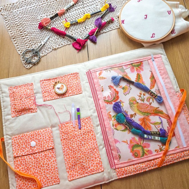 Vue intérieure de la trousse de brodeuse Savannah avec des échevettes de fils glissées dans la pochette zippée en vinyle transparent, sur fond de tissu imprimé oiseaux et fleurs, création artisanale Angélique La Fée En Fil.