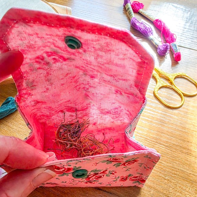Vue plongeante de l'intérieur d'une mini poubelle à fils pliable ouverte modèle Romane, montrant sa doublure rose remplie de chutes de fils, posée sur une table en bois avec ciseaux dorés et échevettes en arrière-plan, création artisanale Angélique La Fée