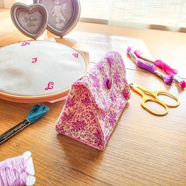 Vue de profil d'une mini poubelle à fils de table pliante fermée modèle Samba en tissu coton à motifs floraux rose et violet, posée sur une table en bois avec un tambour à broder, des ciseaux dorés et des échevettes, création artisanale Angélique La Fée E