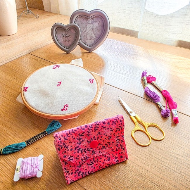 Vue de face d'une mini poubelle à fils pliable fermée pour broderie et point de croix, modèle Sandy Rose en tissu rose aux motifs de feuillage violet avec rabat et bouton-pression rose, posée sur une table en bois à côté d'un tambour à broder, de ciseaux 