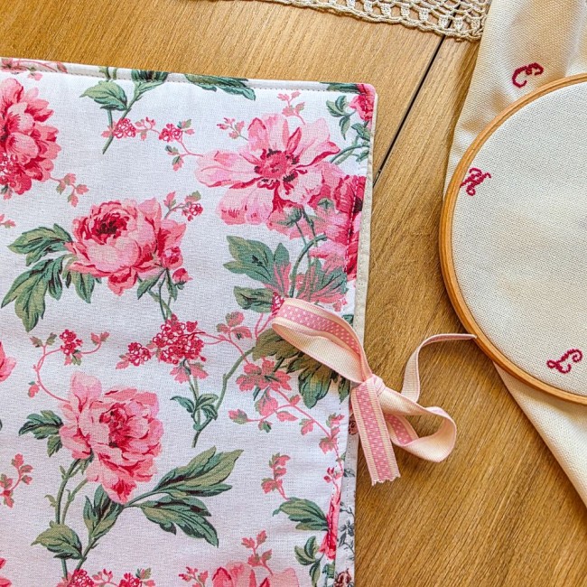 organisateur broderie-sofia-fleurs-roses-vintage Gros plan sur l'extérieur de la trousse de brodeuse Sofia, en tissu vintage à motifs de grandes fleurs roses et pivoines, fermée par un ruban rose poudré. Un tambour à broder est visible en arrière-plan. Création Angélique La Fée En Fil
