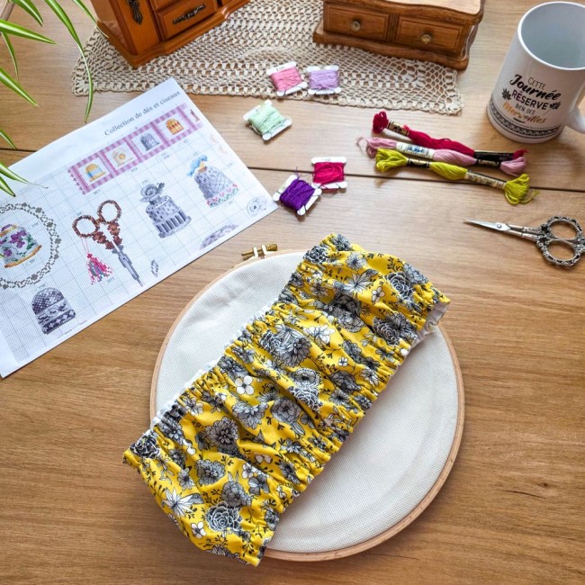 Grime guard modèle Julie en tissu jaune imprimé floral plissé, posé au centre d'un tambour à broder rond en bois avec sa toile à broder sur un bureau d'atelier par Angélique La Fée en Fil.