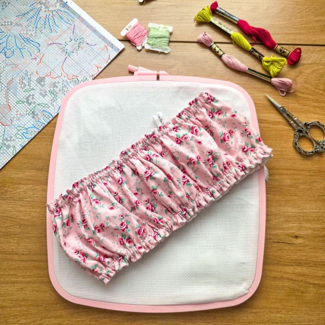 grime-guard-cadre-nurge-point-de-croix-romane-angelique-la-fee-en-fil Vue d'ensemble d'un grime guard housse protège-toile modèle Romane posé en travers sur un cadre à broder rectangulaire rose, en tissu rose aux motifs bucoliques de fleurs et roses avec étiquette de marque, entouré d'une grille de broderie, échevettes et c