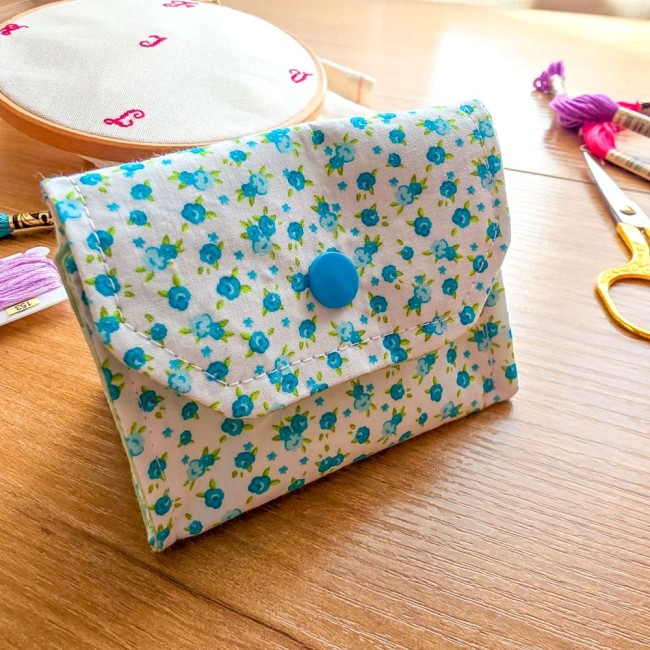 poubelle-a-fils-mini-pochon-broderie-myosotis-vue-face-angelique-la-fee-en-fil Gros plan de face d'une mini poubelle à fils pliable en tissu écru imprimé de petites fleurs de myosotis bleues avec bouton-pression bleu turquoise, posée sur une table en bois clair à côté d'un tambour à broder, échevettes et ciseaux dorés, création arti