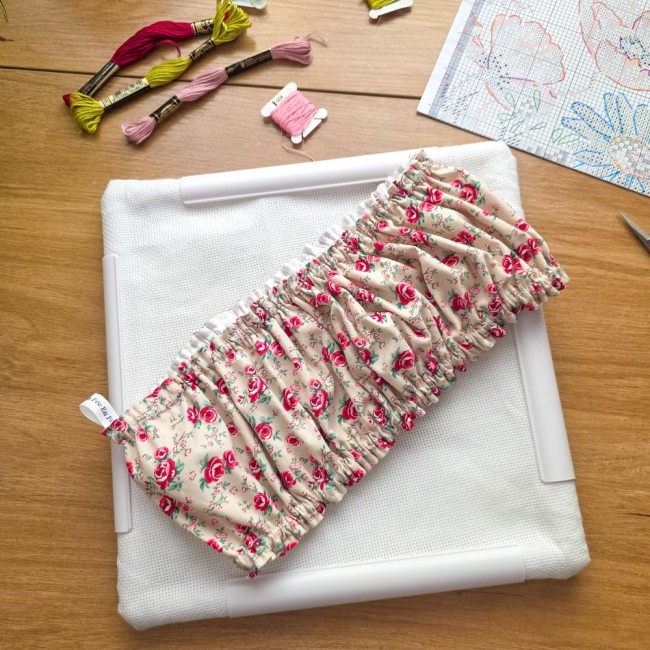 Vue de dessus d'un grime guard housse protège-toile modèle Vanille posé en travers sur un cadre Q-Snap carré blanc avec toile Aïda, en tissu écru vanille aux motifs bucoliques de roses roses et rouges avec étiquette de marque, entouré d'échevettes et d'un