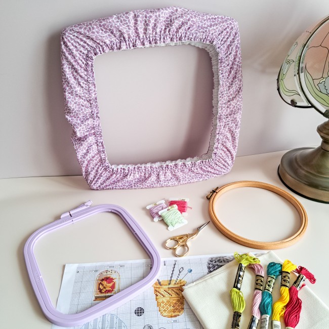 Grime Guard en tissu mauve Lavande de la collection Lumière Végétale sur un cadre carré. Composition créative incluant un petit tambour rond et un nurge, des ciseaux de broderie fins et des fils moulinés aux teintes printanières.