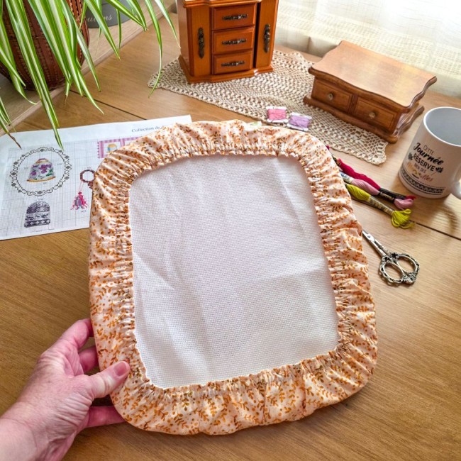 grime-guard-broderie-sous-bois-dore-tissu-beige-feuillage-main-maintien-qsnap-angelique-la-fee-en-fil317 Main tenant un grime guard modèle Sous-Bois Doré en tissu beige à motifs de feuillage ocre houssant un métier Q-Snap, posé sur un bureau de broderie par Angélique La Fée en Fil.