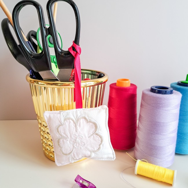 bijou-ciseaux-brodeuse-coussinet-fleur-blanche-ruban-rose-fils-bobines-pot-dore-atelier-angelique-la-fee-en-fil Bijou de ciseaux pour brodeuse en forme de coussinet blanc brodé d’une fleur, suspendu à un pot doré contenant des ciseaux, entouré de bobines de fil rouge, violet, bleu et jaune, création artisanale Angélique La Fée En Fil.
