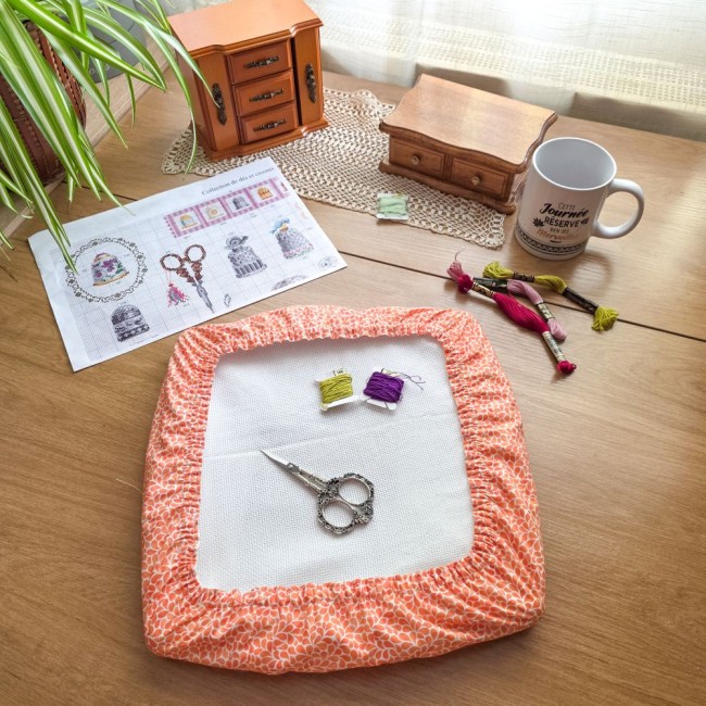 grime-guard-protection-toile-a-broder-cadre-nurge-rectangulaire-point-de-croix-mandarine-orange-angelique-la-fee-en-fil Vue d'ensemble d'un grime guard housse protège-toile modèle Mandarine en tissu coton orange imprimé de motifs gouttelettes blanches, ajusté sur un cadre rectangulaire de type Nurge, avec grille de broderie, ciseaux vintage, cartes à fils et mug sur une ta